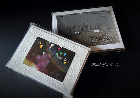 Two open greeting cards with white decorative borders displayed on dark background, showing colorful thank you designs and metallic elements