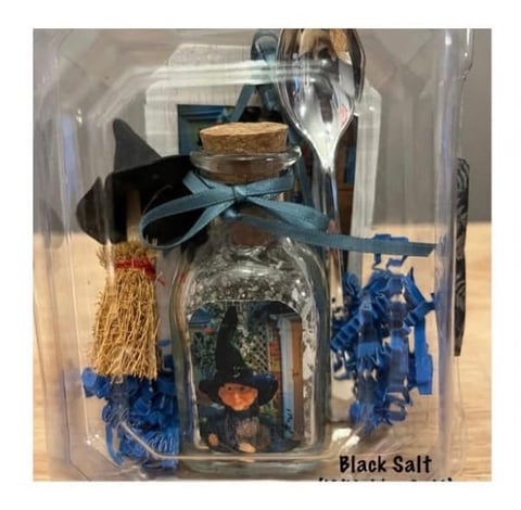 Black salt in decorative glass bottle with cork stopper and blue ribbon bow, displayed in clear packaging with tassels